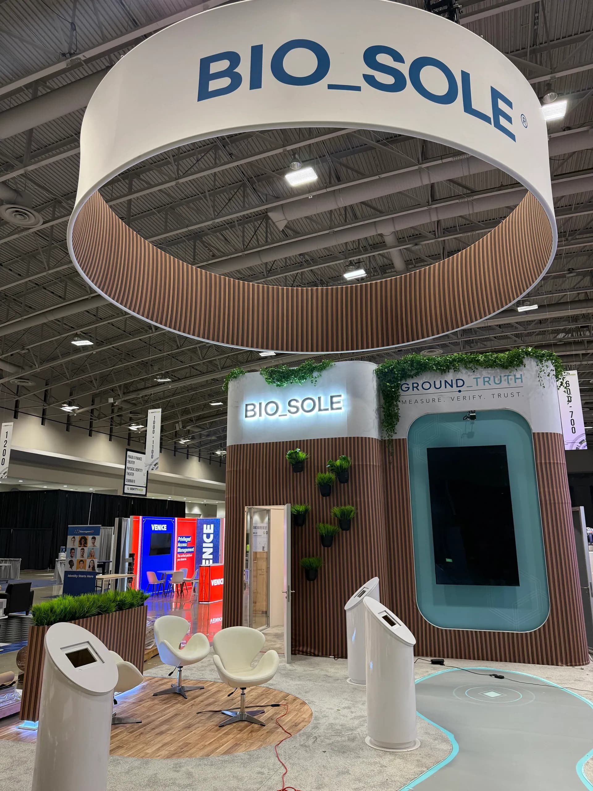 La façade du stand Bio_Sole : une enseigne BIO_SOLE lumineuse et un écran Ground_Truth encastrés dans des lattes de bois garnies de verdure, deux fauteuils blancs et des bornes de vérification autoportantes devant