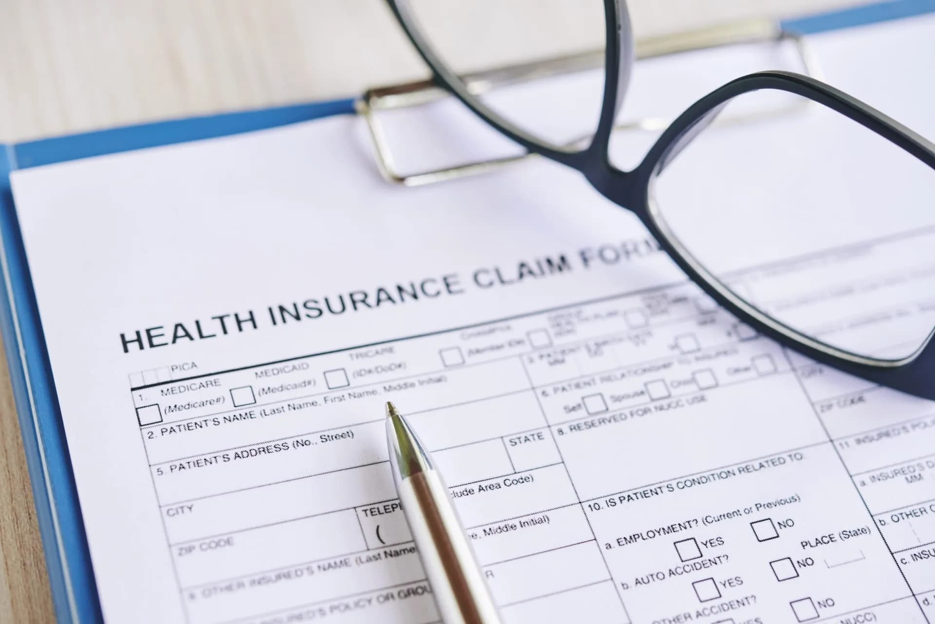 A health insurance claim form on a blue clipboard, reading glasses and a pen resting on top of it